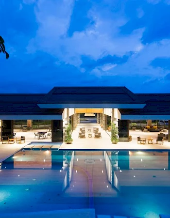 A modern building with a large, illuminated pool in front, surrounded by chairs and trees, set against an evening sky.