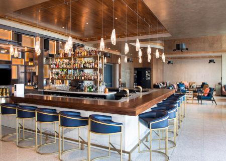 The image shows a modern bar with a wooden countertop, blue chairs, pendant lights, and shelves stocked with bottles, in a stylish lounge area.
