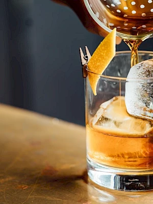 A hand pours a drink into a glass with ice and a lemon peel garnish on a table.