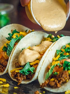 Three tacos with ground meat, cheese, tomatoes, greens, and sauce being poured on top on a glass plate.