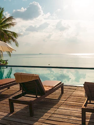 A serene beachside pool scene with lounge chairs, umbrellas, palm trees, and a calm ocean under a bright sky.