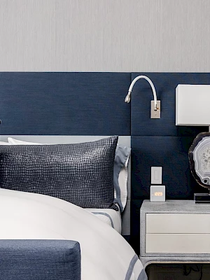 A modern bedroom features a bed with blue and white linens, a blue headboard, beside a nightstand with a unique lamp and a wall lamp.