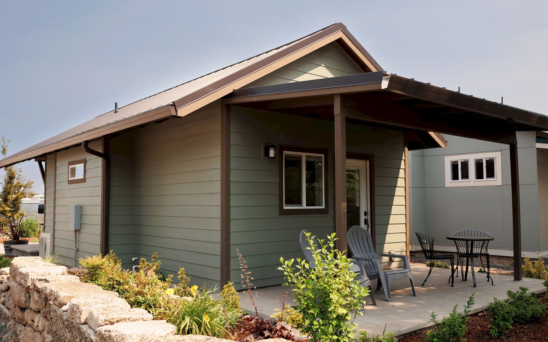 A small, cozy house with a covered porch, outdoor chairs, table, and surrounding plants under a clear sky.