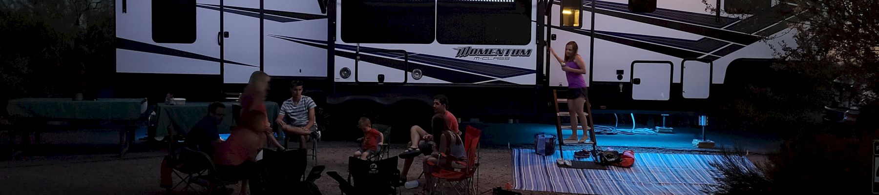 A group of people sit outside an illuminated RV at night, with one person standing near the RV on an elevated platform.