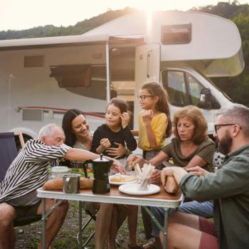 A family enjoys a meal outdoors beside a camper, surrounded by nature. They appear happy and relaxed, sharing food and smiles.