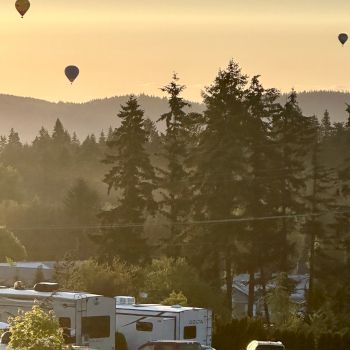 Hot air balloons float in the sky above a forested area with RVs and cars below during a serene sunset.