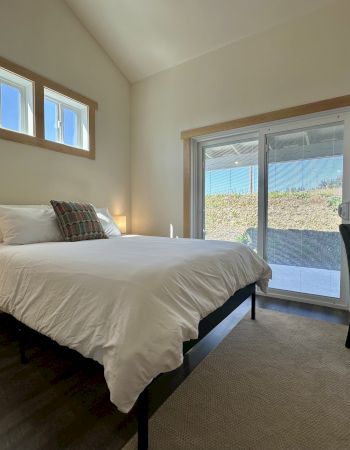 A minimalistic bedroom with a bed, white bedding, two small lamps, chair, and sliding glass doors leading outside.