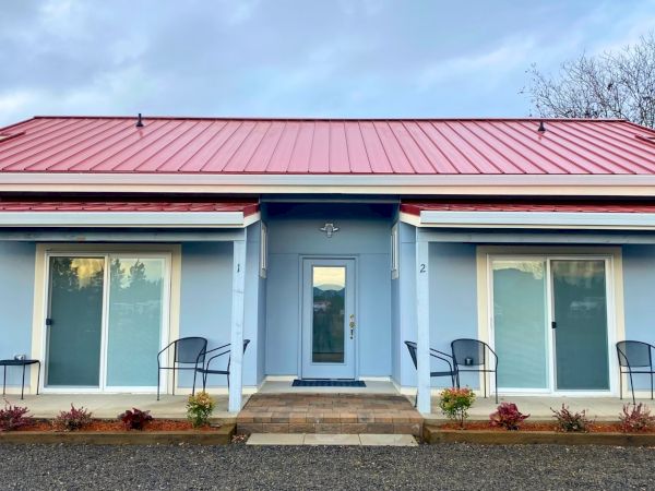 A blue building with a red roof features two sliding glass doors and small seating areas, surrounded by plants at the front.