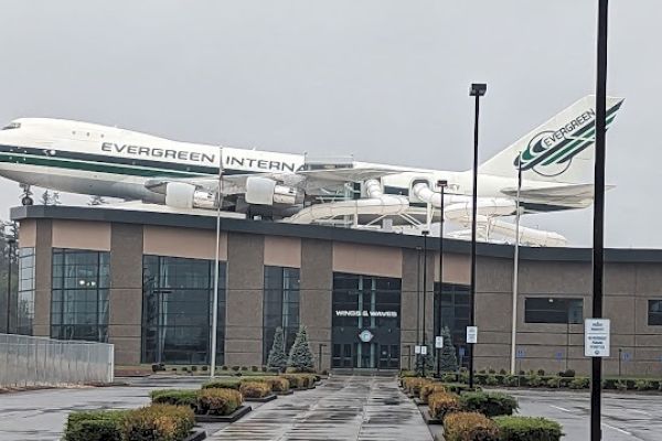 A Boeing 747 with "EVERGREEN" written on it is mounted atop a building. The setting is overcast and features a parking area in front.
