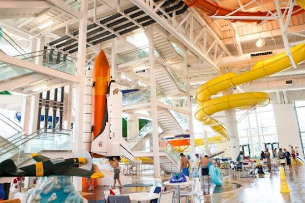 This image shows an indoor waterpark with a space shuttle model, a yellow spiral slide, and a plane model, creating a themed environment.