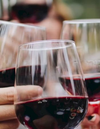 A group of people clinking glasses of red wine in a celebratory setting, with a blurred background suggesting a social gathering.