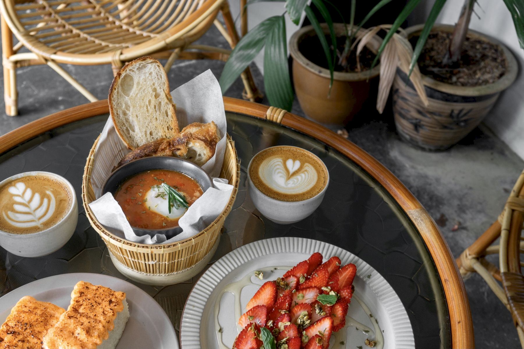 A table with coffee, bread, a dish with tomato and bread slices, and strawberry-topped dessert. There's also a plant in the corner.