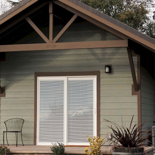 A small house with sliding doors, a chair on the porch, and plants in the garden.