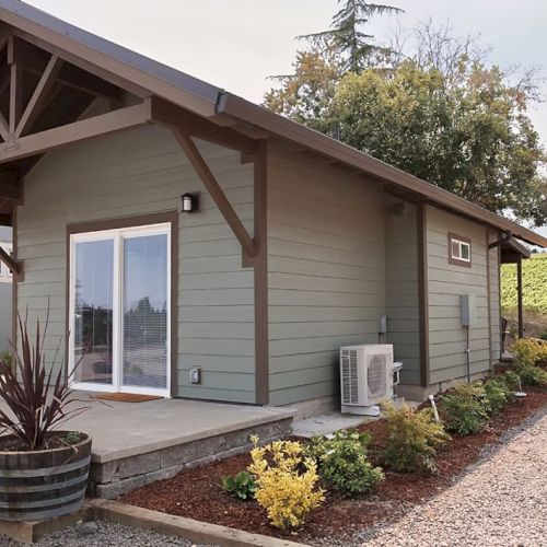 A small, modern cabin with sliding doors, surrounded by plants and gravel, and a vineyard visible in the background.