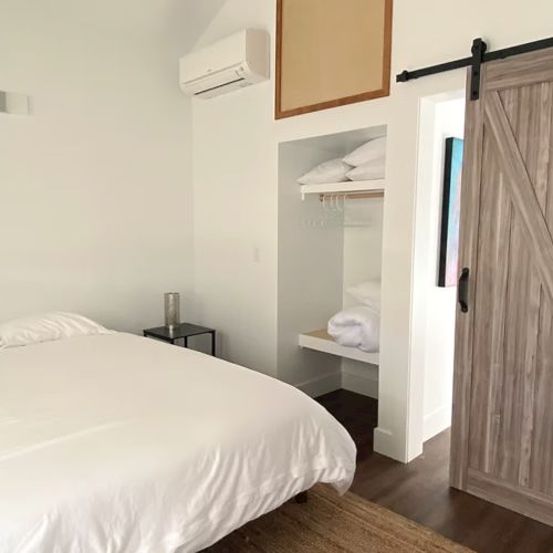 A minimalist bedroom with a white bed, sliding barn door, open closet with shelves and a wall-mounted air conditioner unit.