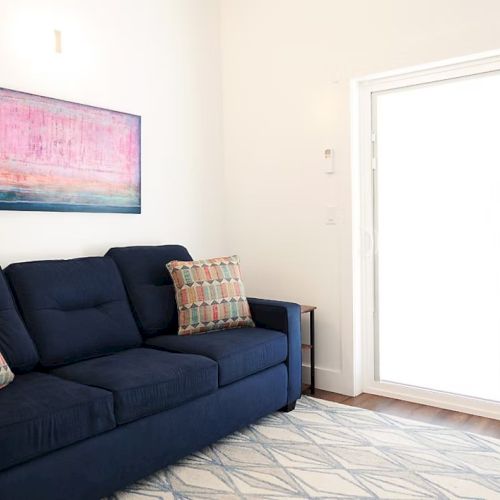 The image shows a room with a dark blue sofa, patterned cushions, abstract art on the wall, and a large sliding glass door providing natural light.