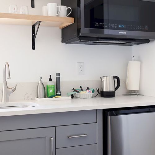 A compact kitchen with a sink, overhead microwave, kettle, and mini fridge; shelves hold mugs and glasses, and a paper towel and soap are visible.