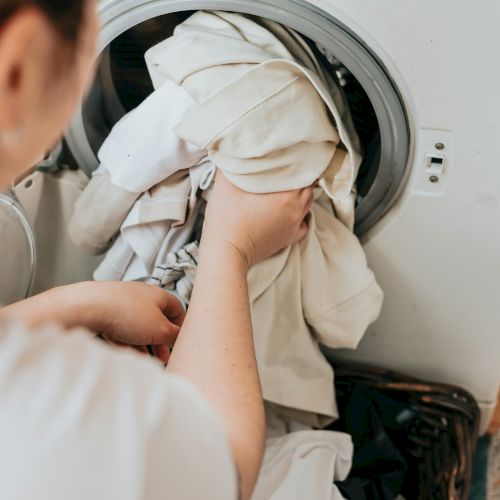 A person is loading or unloading clothes from a washing machine, with several light-colored garments visible in the machine.