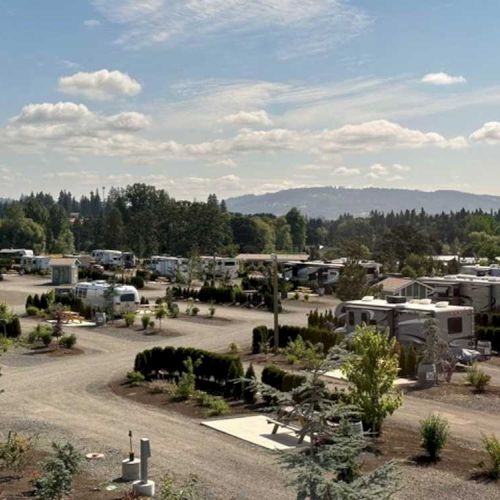 A campground with numerous RVs is set against a landscape of trees and distant hills under a partly cloudy sky.