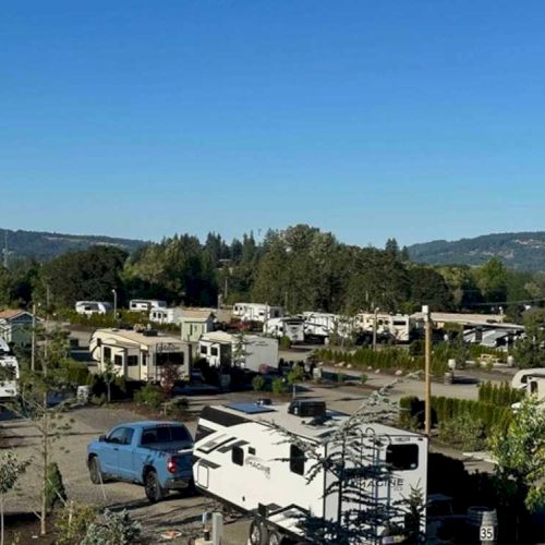 The image shows a campground with numerous RVs and trailers parked, surrounded by trees and hills under a clear blue sky.