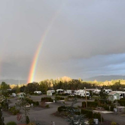 A double rainbow arcs over an RV park under a cloudy sky, with trees and mountains in the background.