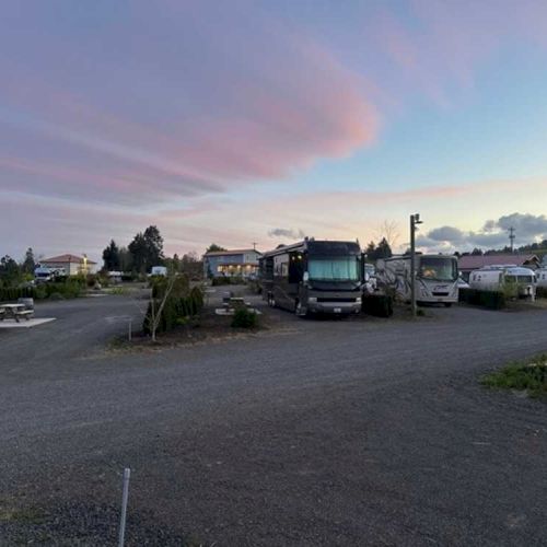 The image shows a campground with several RVs parked under a colorful sky at dusk, surrounded by trees and open space.