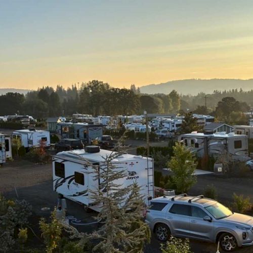 The image shows a campground with numerous RVs and cars parked among trees, set against a misty, scenic backdrop at sunrise or sunset.