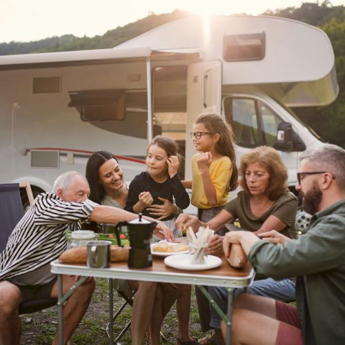 A group of people are enjoying a meal together outdoors beside a camper, surrounded by nature at sunset.
