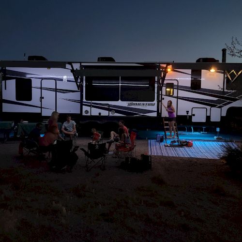 A large RV is parked at dusk, with people gathered around a seating area outside, illuminated by lights.