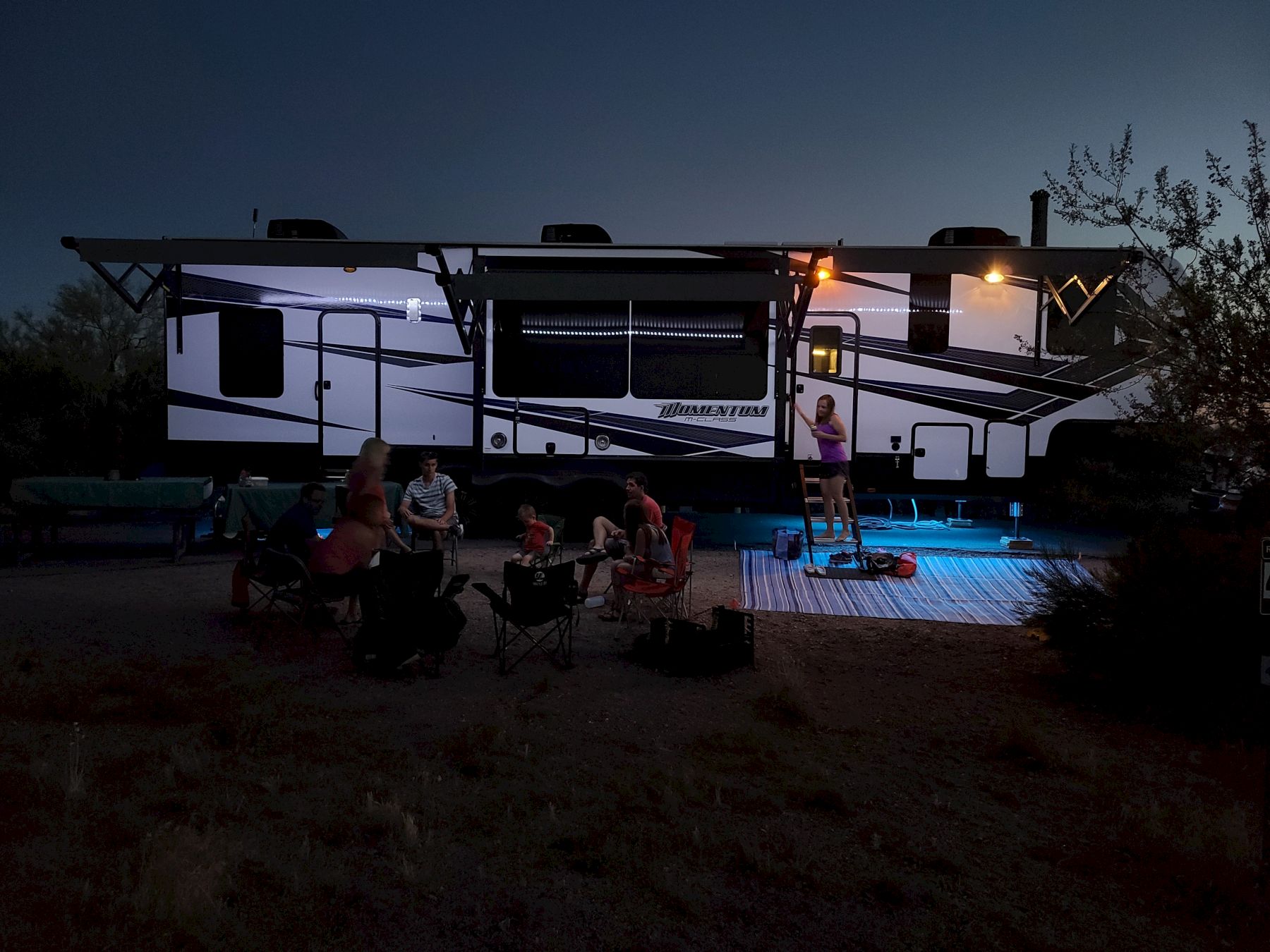 A group of people is gathered outside a large RV at dusk, with one person on a ladder, and soft lights illuminate the scene.