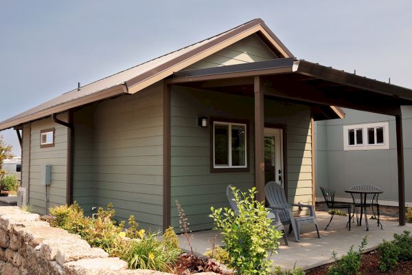 A small house with a covered porch, two patio chairs, a table, and surrounding greenery on a sunny day.