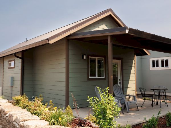 A small house with a covered porch, two patio chairs, a table, and surrounding greenery on a sunny day.